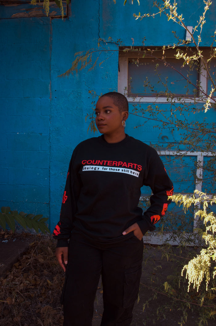 A person with short hair stands outdoors by a blue wall and overgrown plants, wearing a black Counterparts long-sleeve shirt with red and white printed text, gazing thoughtfully to the side.