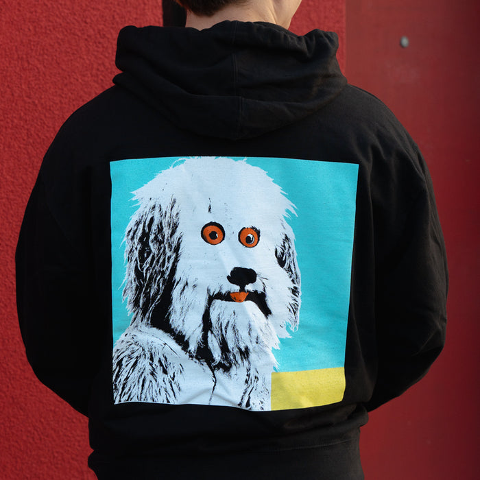 A person wears a black Four Year Strong hoodie featuring a large, colorful graphic of a white shaggy dog with bright orange eyes on a blue and yellow background.
