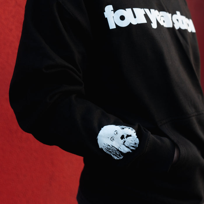 A person wears a Four Year Strong hoodie featuring four year strong printed on the chest and a white dogs head graphic on the sleeve, standing in front of a red wall.
