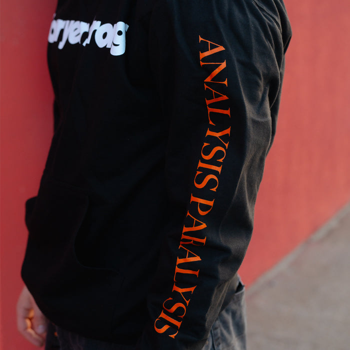 A person wearing a Four Year Strong hoodie with ANALYSIS PARALYSIS in orange on the sleeve and part of a white logo on the chest, standing against a red wall.