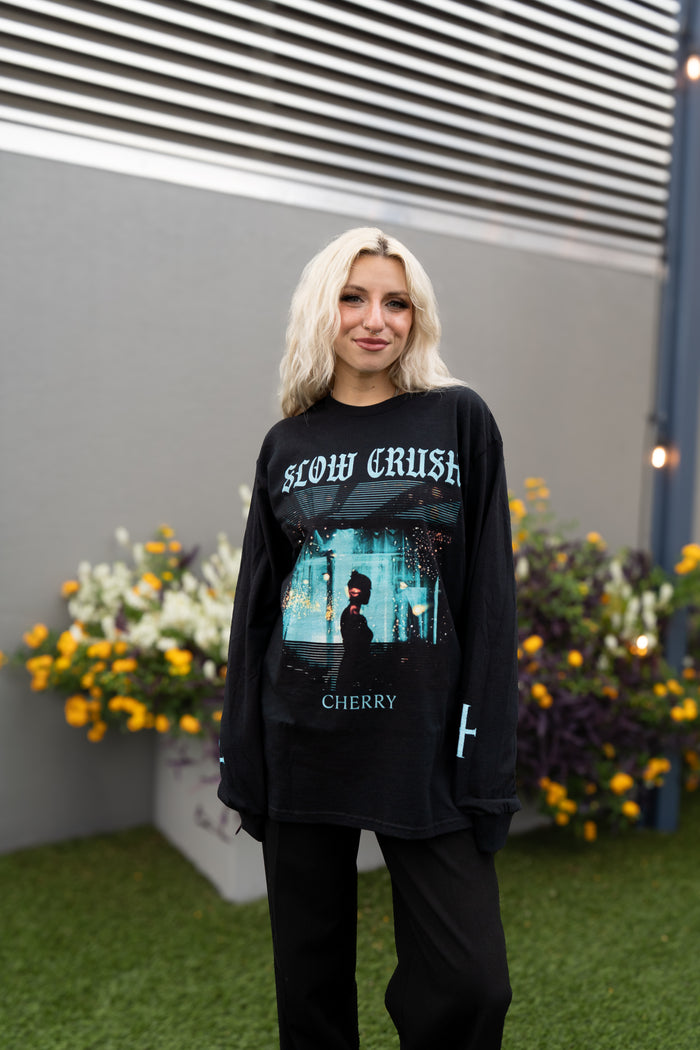 A person with shoulder-length blond hair stands on grass, wearing a black Slow Crush Cherry print t-shirt and black pants. Planters of yellow and white flowers are visible behind them.