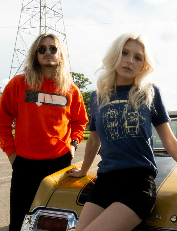 Two people with long blonde hair stand by a yellow vintage car. One wears an orange hoodie and sunglasses, while the other sports a blue DOWNWARD printed shirt and black shorts. A tall metal tower is visible in the background.