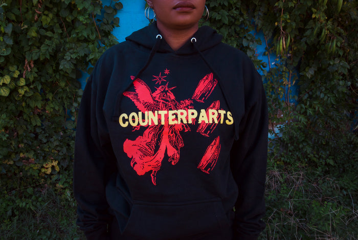 A person wearing a black Counterparts hoodie with bold red graphics and the brand name in large yellow letters stands before a wall of green vines.