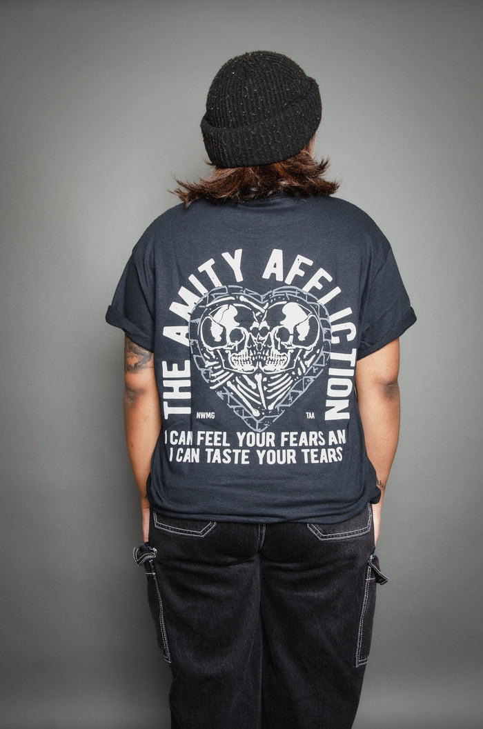 A person wearing a black beanie and The Amity Affliction black T-shirt featuring two skulls on the back stands facing away from the camera against a gray background.