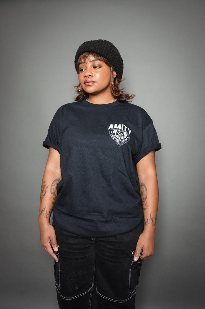 A woman wears a black beanie and a black AMITY t-shirt by The Amity Affliction, standing against a gray background. She has tattoos on her arms and gazes to the side with a neutral expression.
