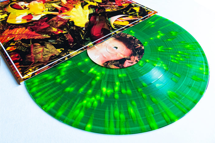 A vibrant green and neon yellow splatter vinyl record by Drug Church partially emerges from a colorful, abstract, nature-themed album cover—all set against white—presenting the striking Hit Your Head vinyl LP presale edition.