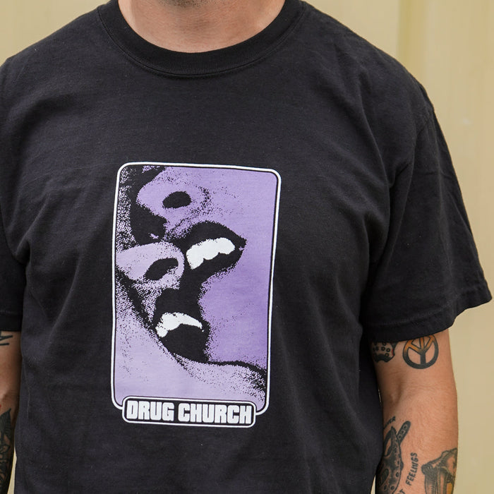 A person with arm tattoos wears a black Drug Church Comfort Colors t-shirt featuring two stylized open mouths on a purple background and the text DRUG CHURCH below.
