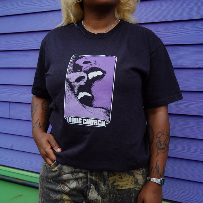 A person with blonde hair wears a black printed Drug Church T-shirt, featuring two faces on a purple background. The style is similar to Comfort Colors. They stand before a purple wall with green trim, one hand in their pocket.