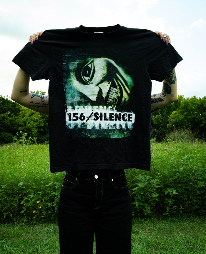 Person holding up a black Comfort Colors T-Shirt with a graphic of a fish head and the text 156/SILENCE, standing outdoors in front of green bushes and trees under a cloudy sky.