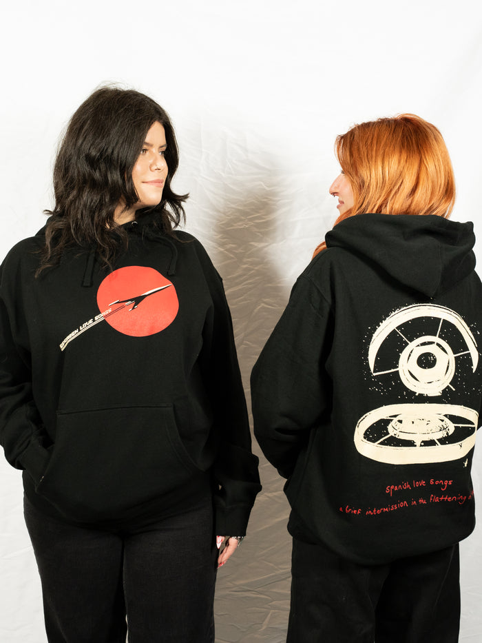 Two women in black printed hoodies from Spanish Love Songs stand facing each other. One hoodie has a red circle graphic with text, while the other features a large white graphic with red writing on a white background.