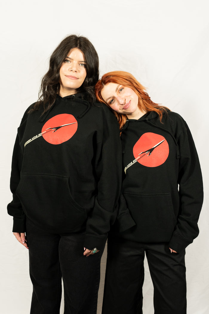 Two women stand side by side against a white background, both wearing printed hoodies from Spanish Love Songs, featuring a large red circle and diagonal yellow lightning bolt graphic. One woman rests her head on the other's shoulder.
