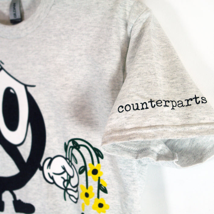 A light gray Counterparts T-shirt featuring counterparts printed on the sleeve and a cartoon hand holding yellow flowers on the front. Ideal for casual, stylish comfort.