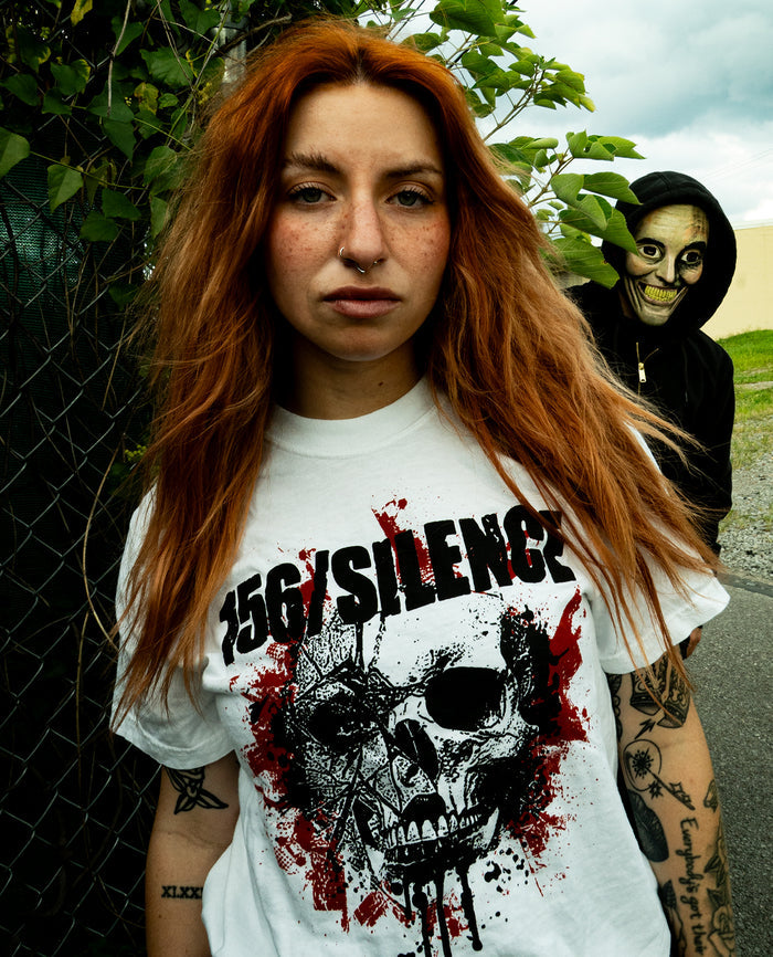 A woman with long red hair and tattoos wears a 156/SILENCE skull graphic shirt made from soft, heavyweight fleece, standing outdoors. Behind her, a person in a hooded jacket and skull mask stares at the camera through green foliage.