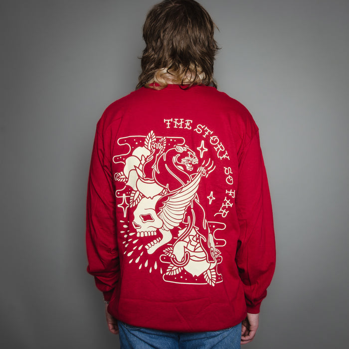 A person with long brown hair wears a red The Story So Far Gildan Heavy Cotton long sleeve shirt with a large white winged skull, roses graphic, and The Story So Far text on the back, standing against a plain gray background.