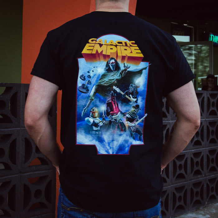 Someone outdoors near a wall with geometric cutouts is wearing a black Galactic Empire Heavy Tee featuring a vibrant graphic of stylized Star Wars characters and ships on the back.