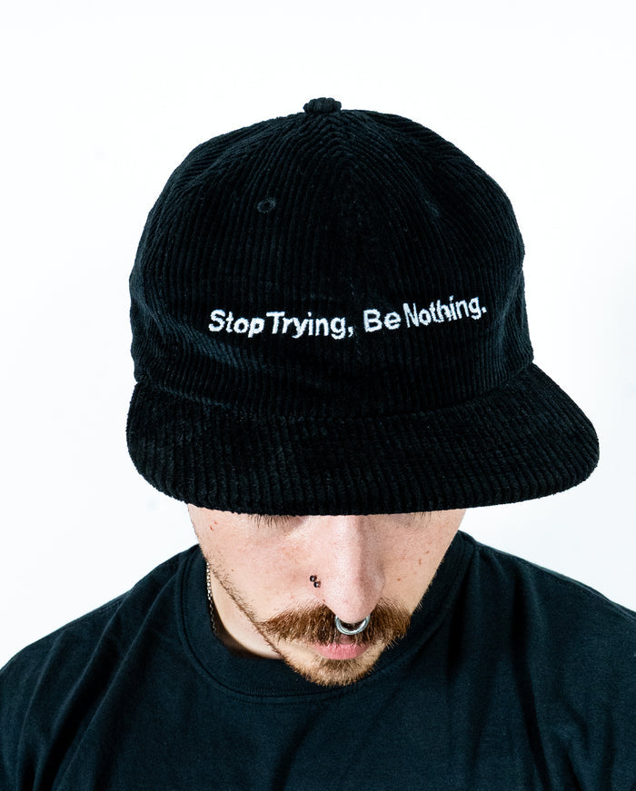 A person with facial piercings and a mustache looks down, wearing a black corduroy hat from Class Caps that says Stop Trying, Be Nothing. The background is plain white.
