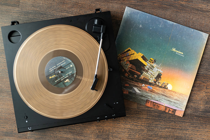 A transparent brown vinyl from Samiam’s 3rd pressing spins on a black turntable, while its cover—showing a surreal, colorful seascape with containers and a sunset—lies nearby on the wooden surface.