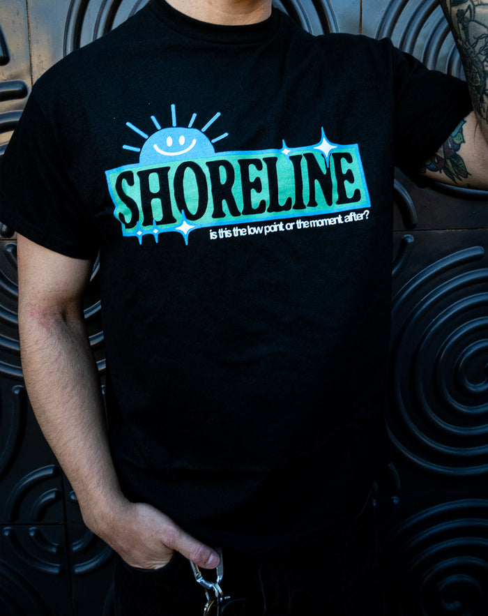 Someone in a black Shoreline Gildan shirt with a blue and green SHORELINE graphic, a smiling sun, and the text "is this the low point or the moment after?" stands before a textured black wall.
