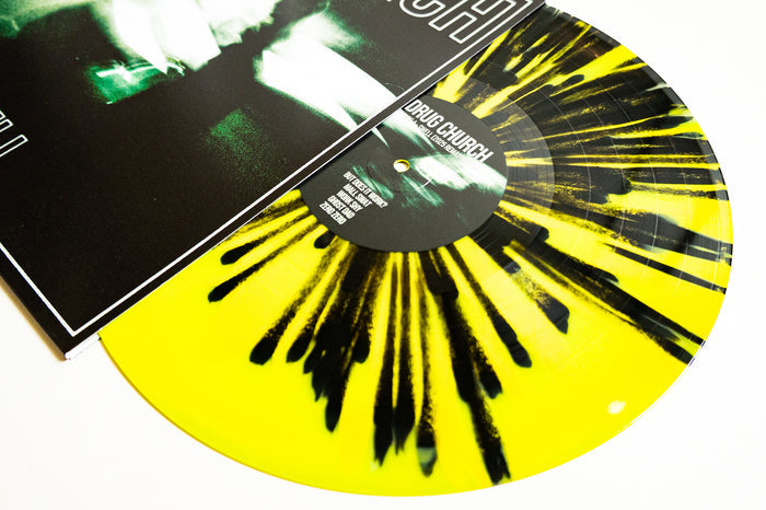 A yellow vinyl LP with black streaks from Drug Church is shown partially inside its album cover. The Coke Bottle/Canary Yellow pressing stands out against a white background, and the label displays "Drug Church" in white text.