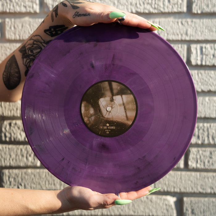 A tattooed person with long green nails holds The Early Novembers Lavender Eco Mix Vinyl by a white brick wall, sunlight highlighting its translucent color and exclusive presale appeal.