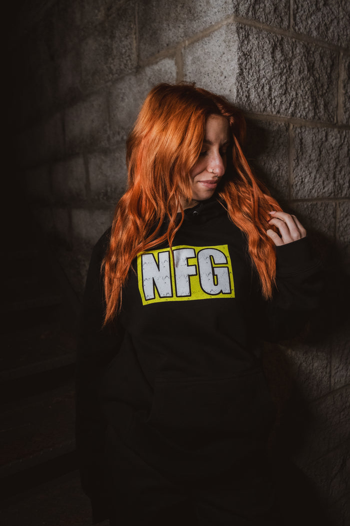 A person with long red hair wearing a black New Found Glory hoodie with bold yellow "NFG" lettering stands against a stone wall, looking down and touching their hair.