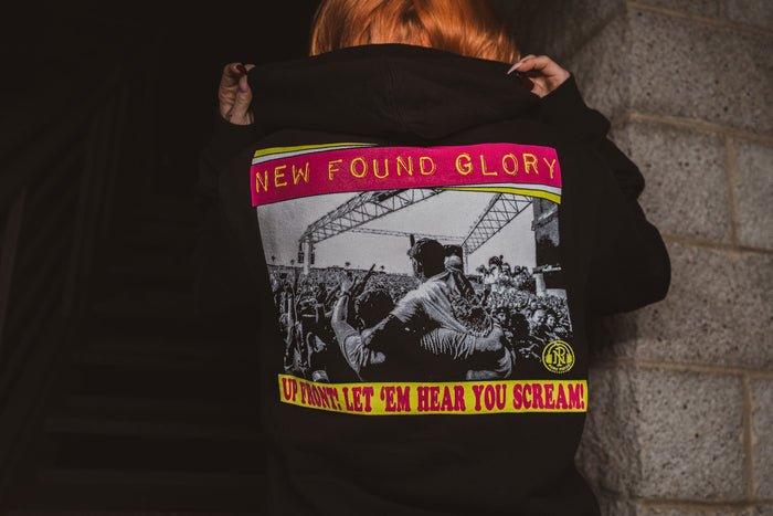 A person with orange hair faces away, pulling up a black New Found Glory hoodie featuring a colorful concert crowd and stage graphic, with yellow and pink text that says, UP FRONT, LET EM HEAR YOU SCREAM!.