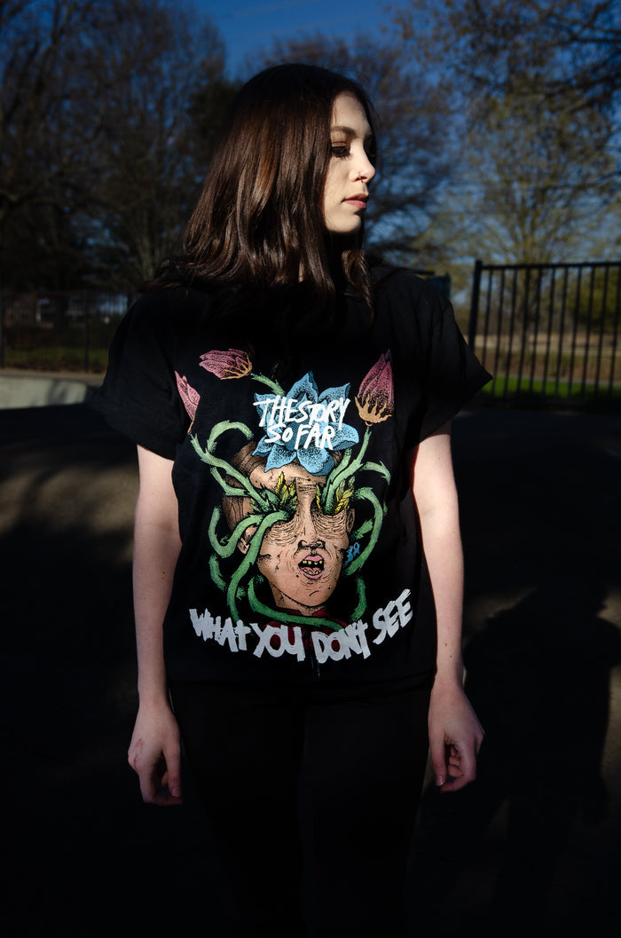 A person with long brown hair stands outdoors by trees and a metal fence, wearing a black The Story So Far band tee featuring colorful screaming head with snakes artwork. The shirt is made from 100% cotton midweight fabric.
